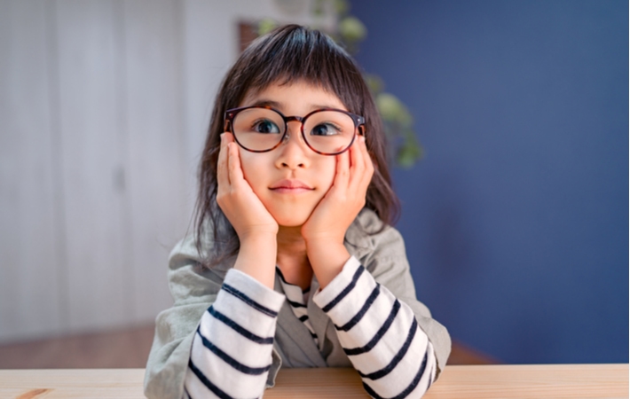 Young girl sits with her elbows at a table holding her head up with her hands. Left eye shows signs of inward turning amblyopia 