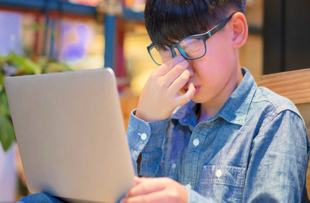 A young boy with glasses rubs his eyes while working on a laptop.