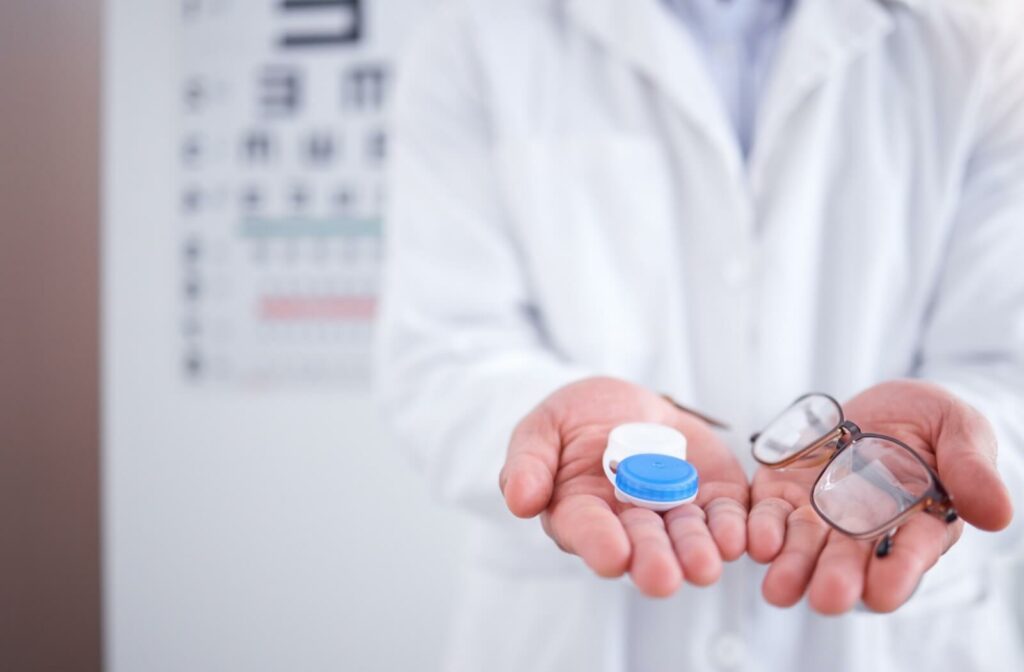 An optometrist holds a pair of glasses and contact lenses in each hand in front of a reading chart.