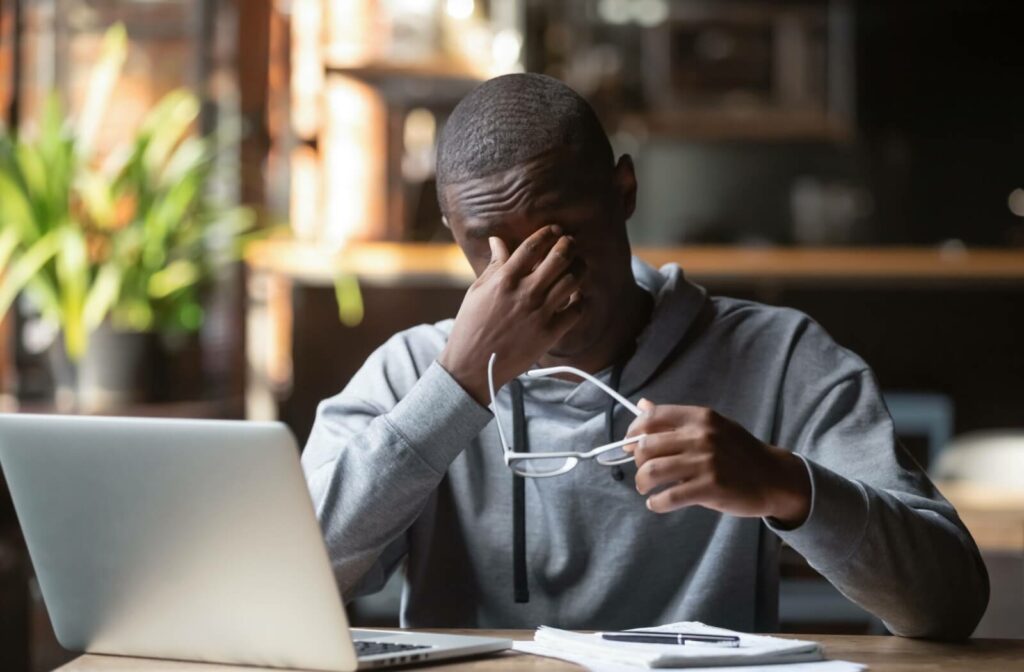 Young adult takes his eye glasses off to rub his eyes while working on his laptop