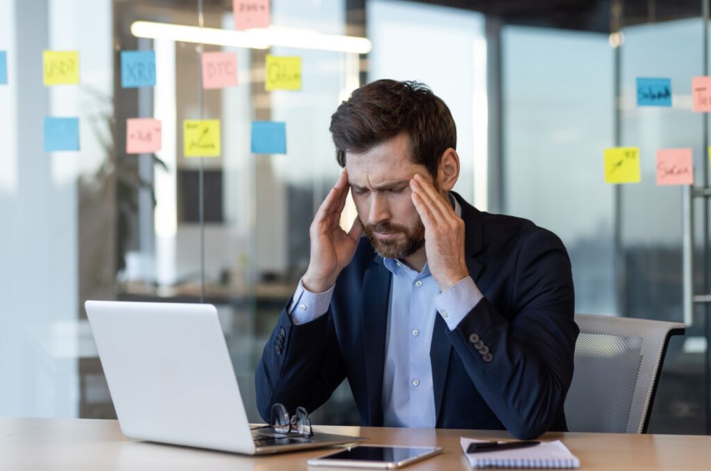 A man sitting at a desk in front of a laptop with eyes clenched and holding his temples from a headache.