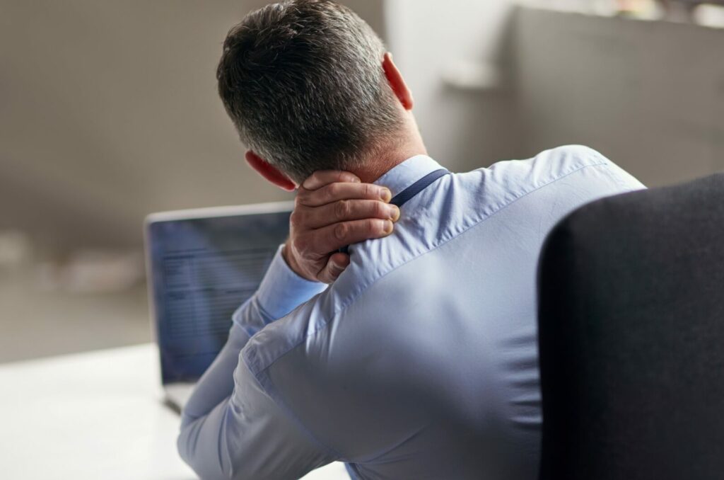 An image of a man from behind sitting in front of a laptop holding his neck with his left hand because of neck pain.