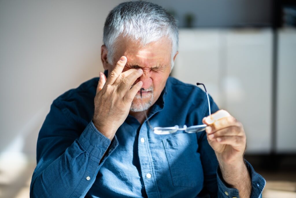 An older man rubbing his eye in discomfort while holding his glasses, showing signs of sudden eye pain.